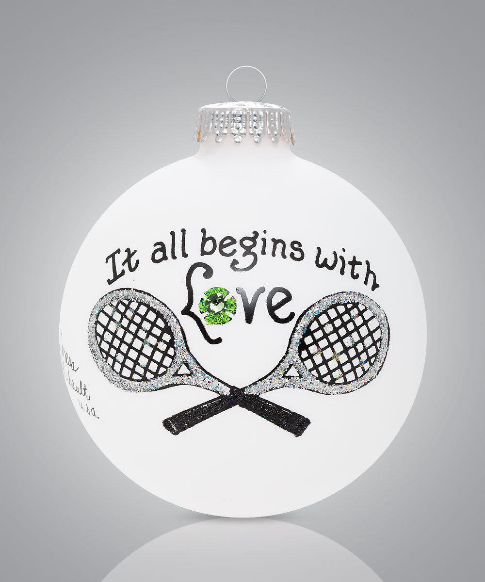 Hand-painted glass tennis ornament with two rackets and the message “It all begins with love”