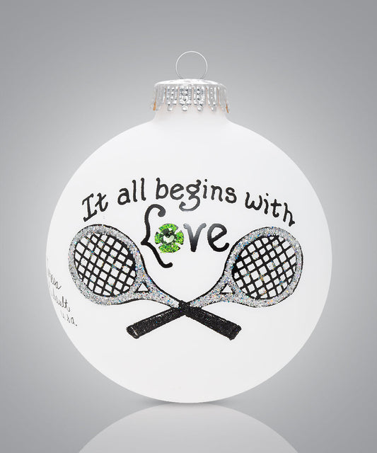 Hand-painted glass tennis ornament with two rackets and the message “It all begins with love”